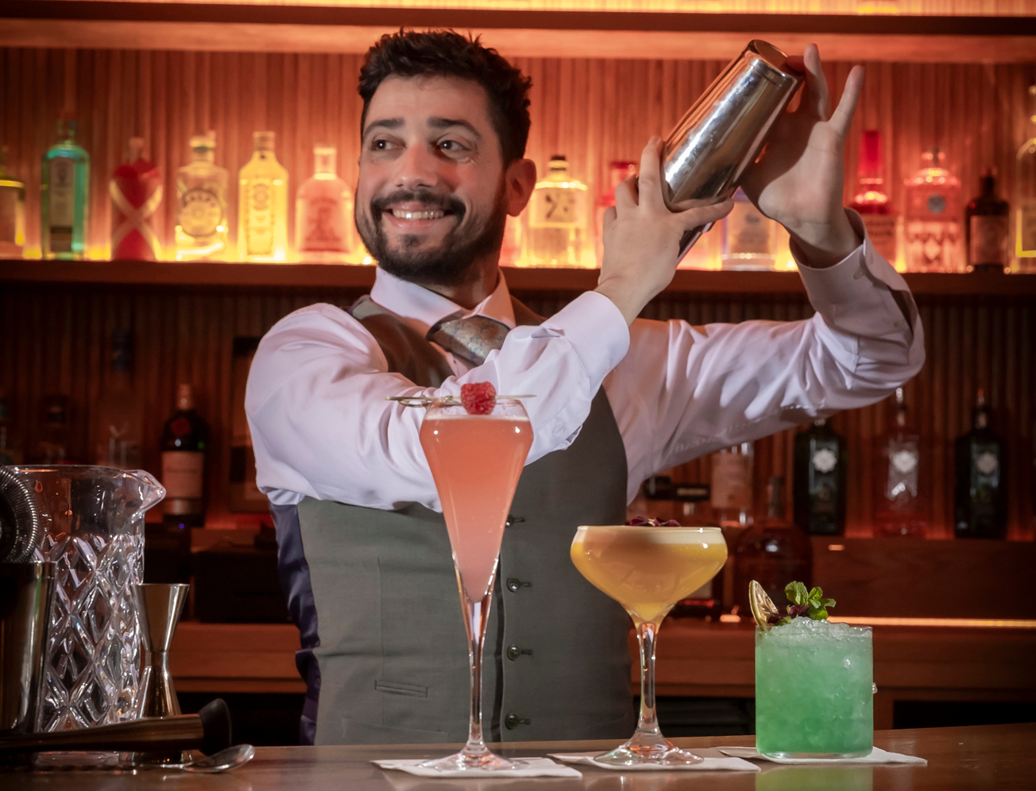 Smiling bartender in a vest and tie shaking a cocktail shaker behind the bar, with three colorful cocktails — pink, yellow, and green — displayed on the counter in front of him against a backdrop of illuminated liquor bottles.