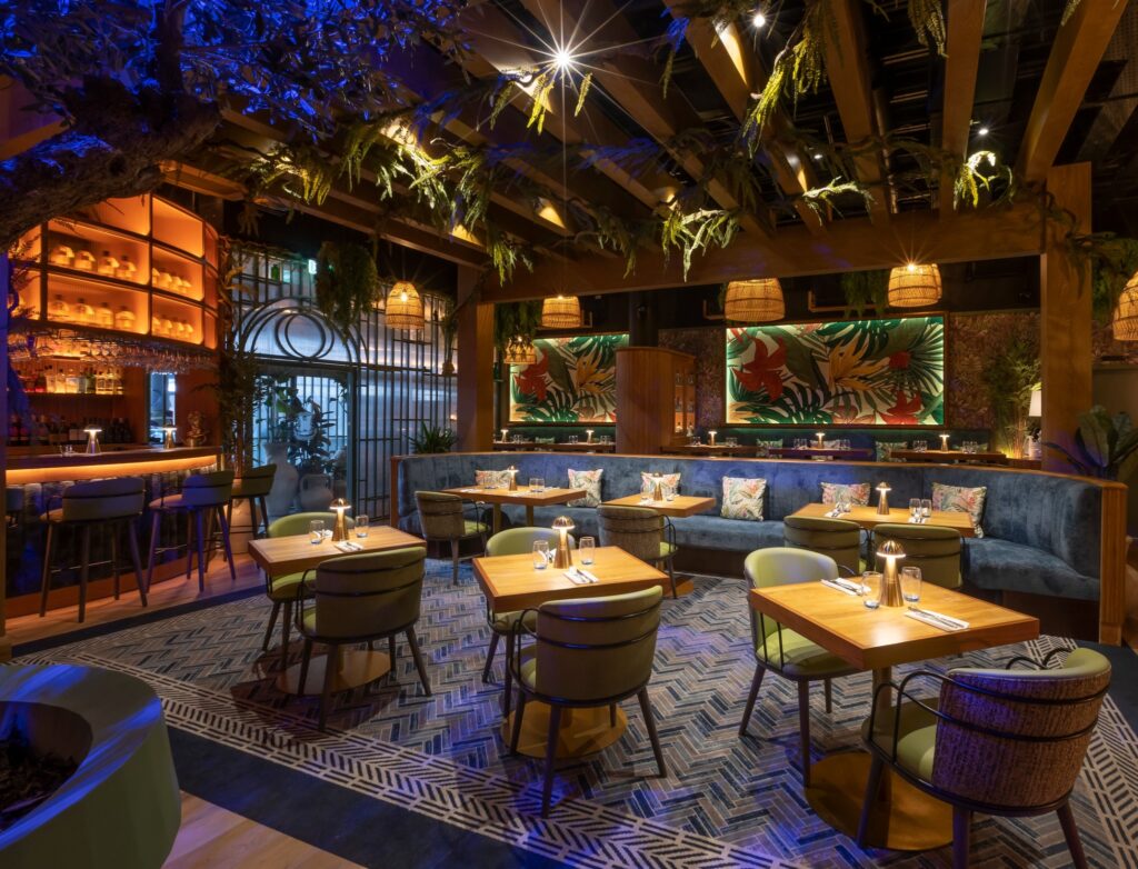 Jekyll restaurant interior with warm lighting, tropical-themed decor, hanging plants, and wooden beams. Tables are neatly set with modern tableware, surrounded by green and blue upholstered seating and a softly illuminated bar area.