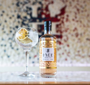 HYDE Gin bottle next to filled gin glass with garnish in front of a flower wall with the hyde logo in the background